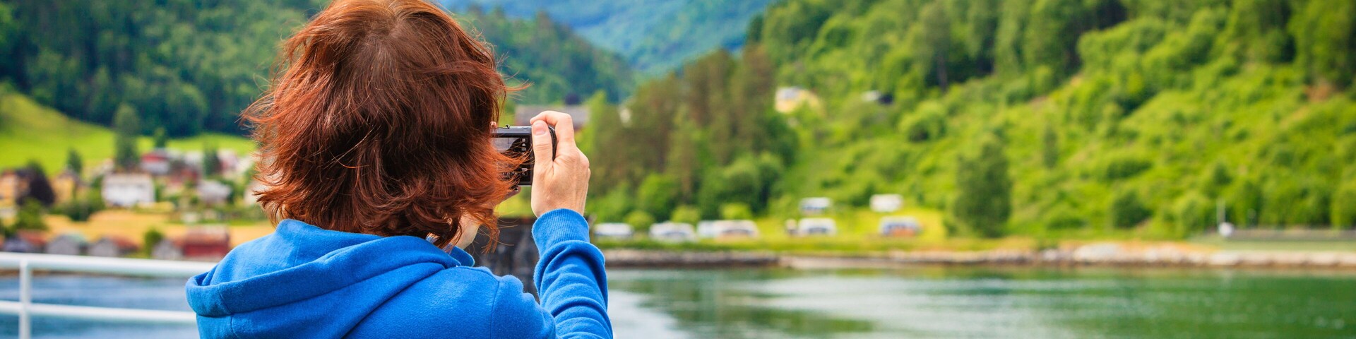 A person in a blue hoodie taking a photo of a lake with mountains in the background.