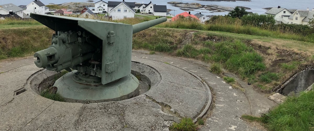 A military cannon on a concrete platform overlooking a coastal village with houses and rocky outcrops.