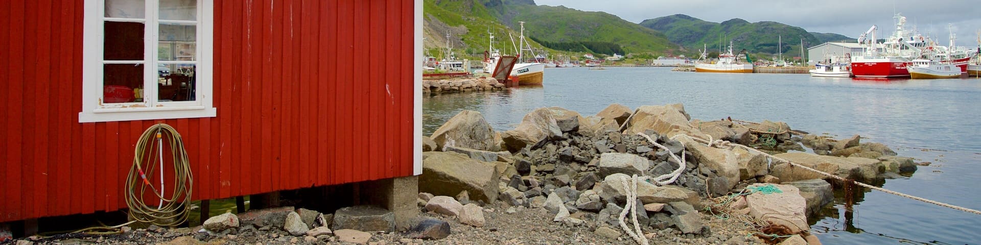 A red wooden building by a rocky shore with boats docked in the background.