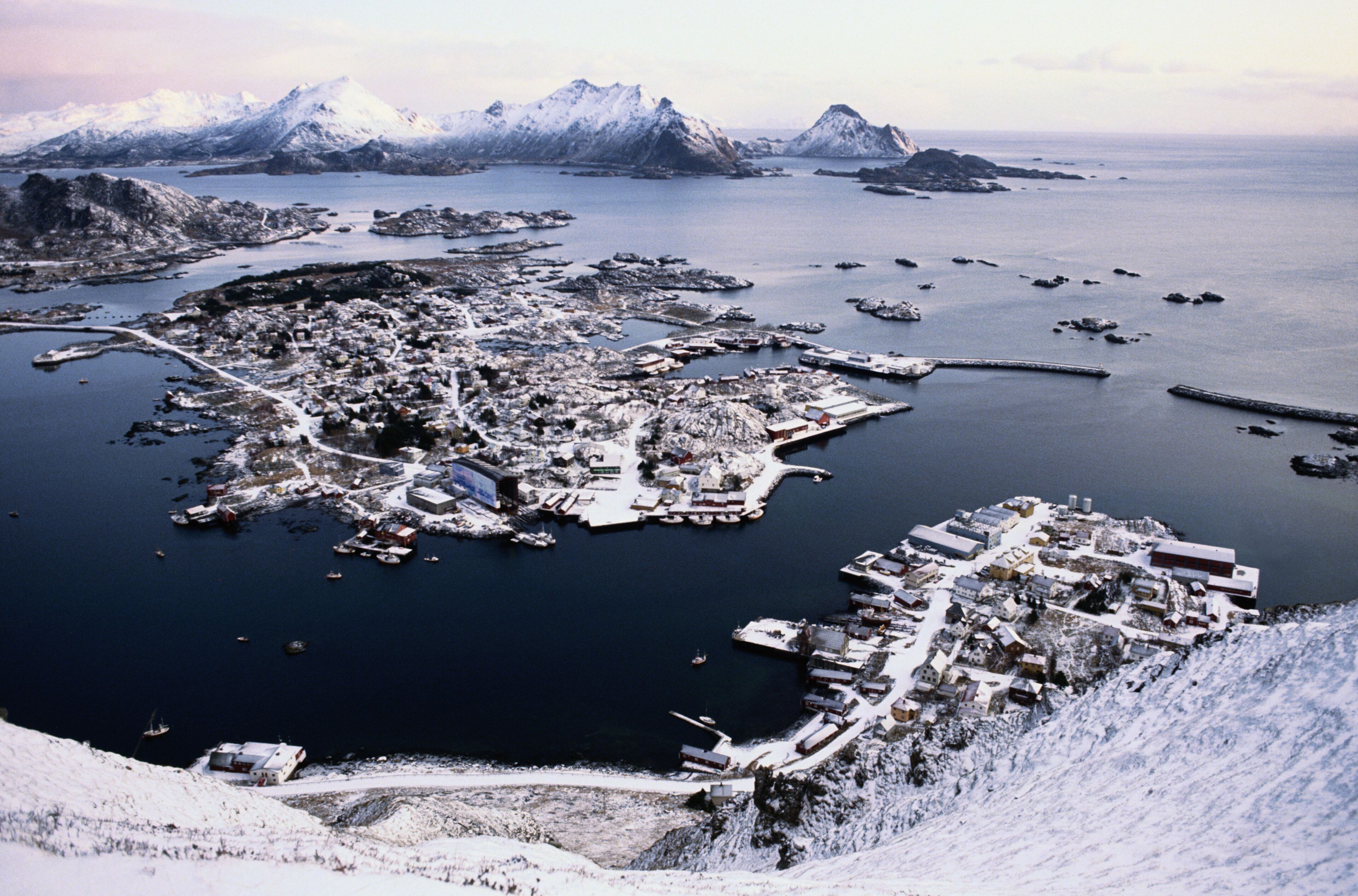 Un pueblo costero cubierto de nieve con un puerto, barcos y montañas al fondo.