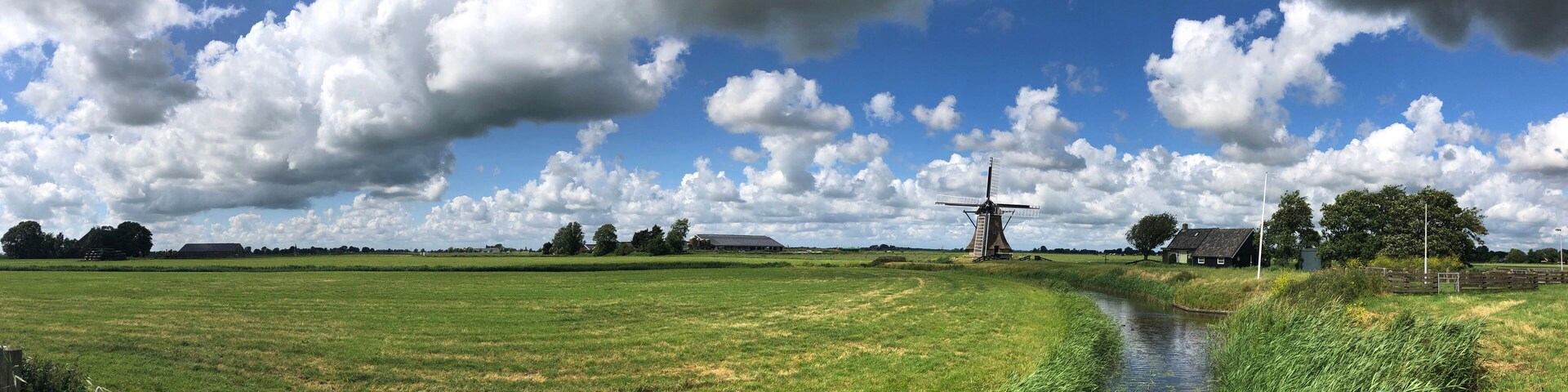 Um moinho de vento, um canal e uma paisagem rural com campos e árvores sob um céu parcialmente nublado.