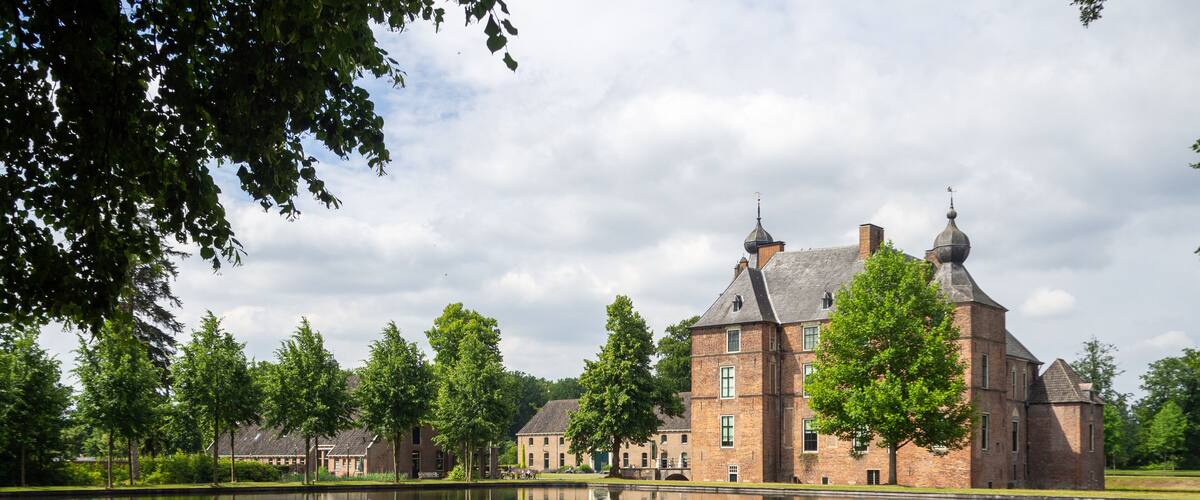 A large brick castle with multiple towers, surrounded by a calm lake and lush greenery.