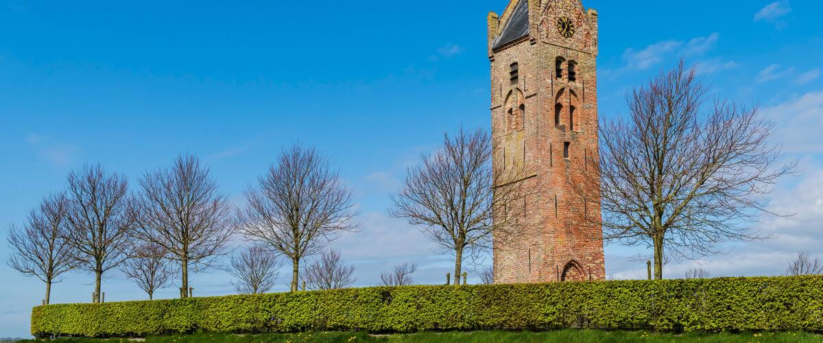 A historic brick tower with a clock, surrounded by a well-maintained hedge and a grassy field with trees.