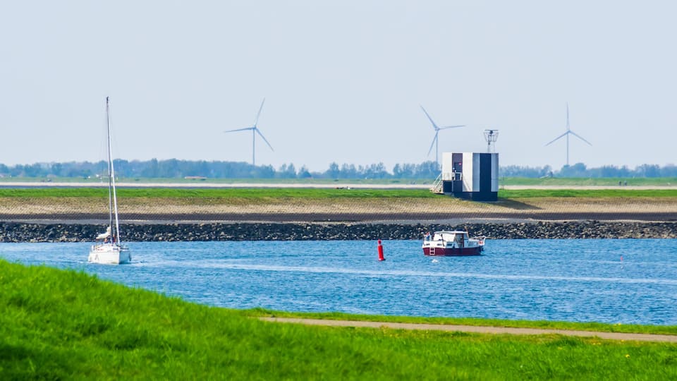 A river with boats, wind turbines, and a grassy bank.