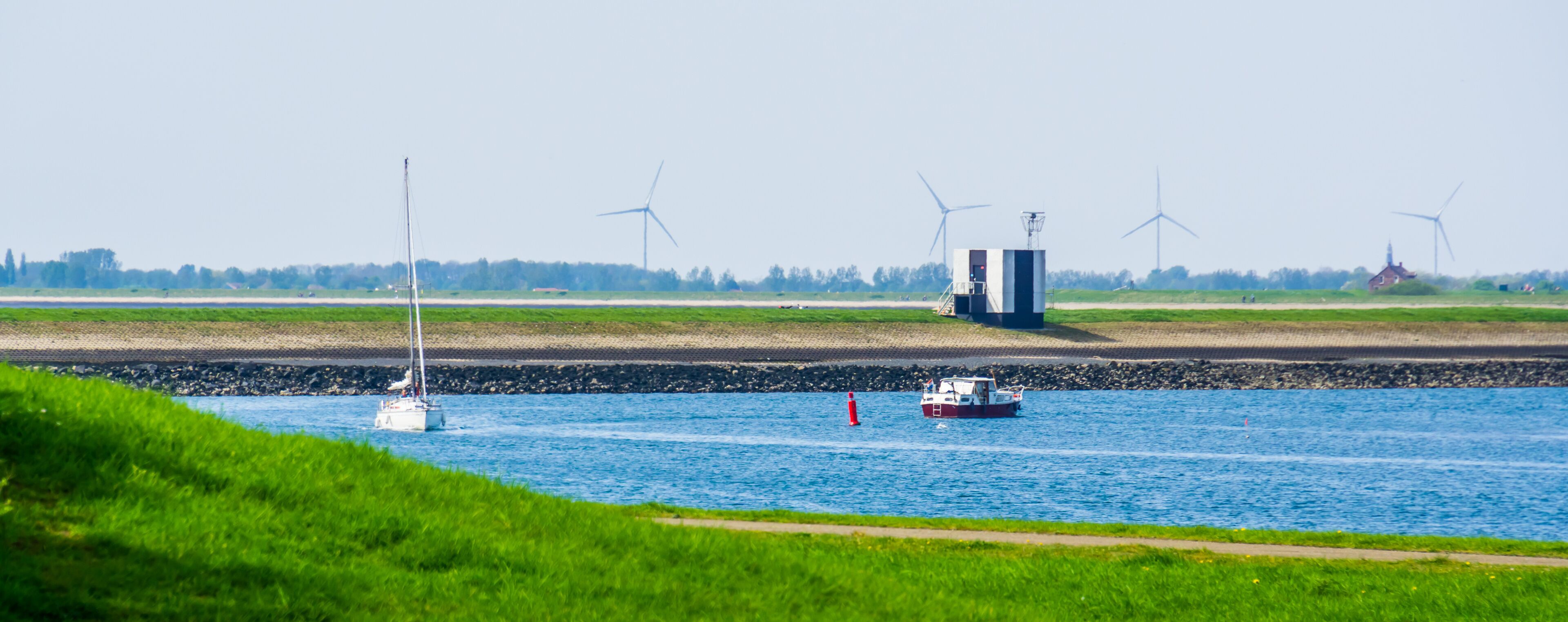 A river with boats, wind turbines, and a grassy bank.