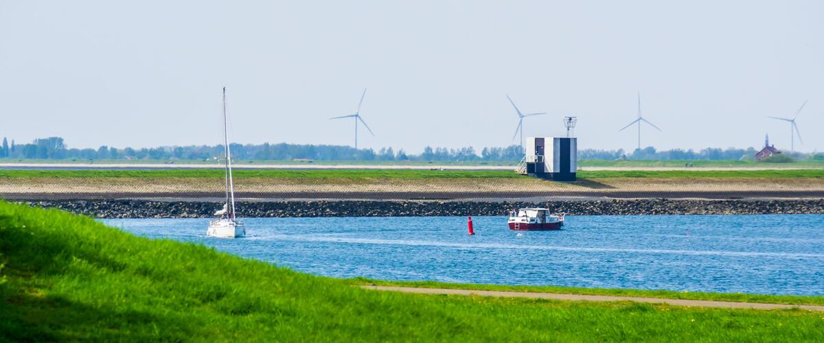 A river with boats, wind turbines, and a grassy bank.
