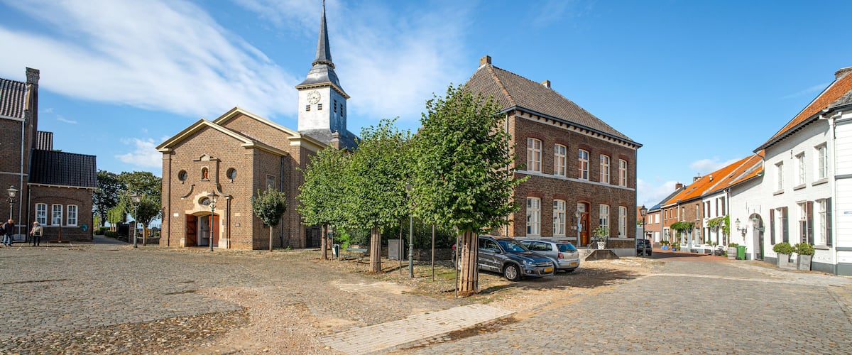 A cobblestone street with historic buildings, a church with a tall steeple, and parked cars.
