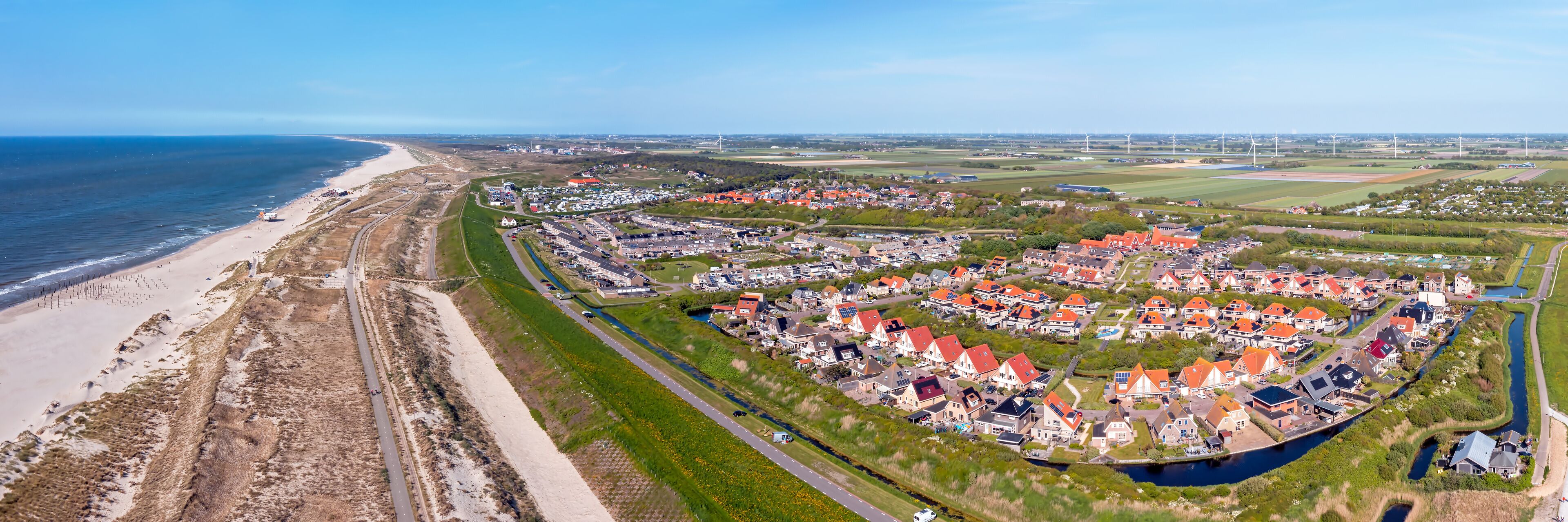 An aerial view of a coastal town with a beach, houses, and wind turbines.