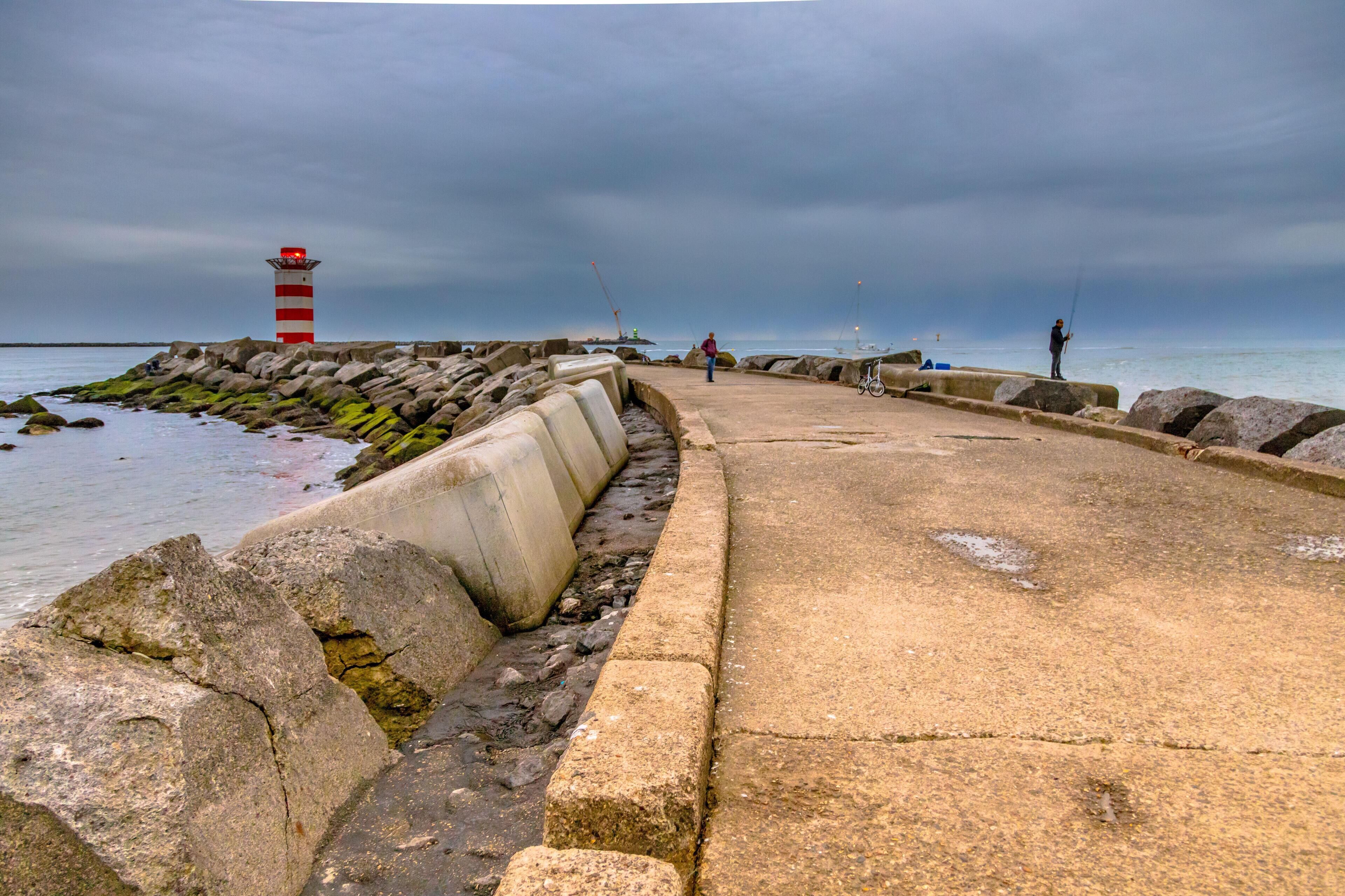 A coastal walkway with a lighthouse, rocky shore, and fishing activity.