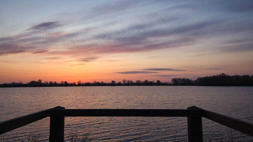 Horizontal view on a silhouette of a wooden railing with a beautiful sunrise, clouds and water at the background. Early morning in spring at “De Zandmeren” in Kerkdriel, Netherlands