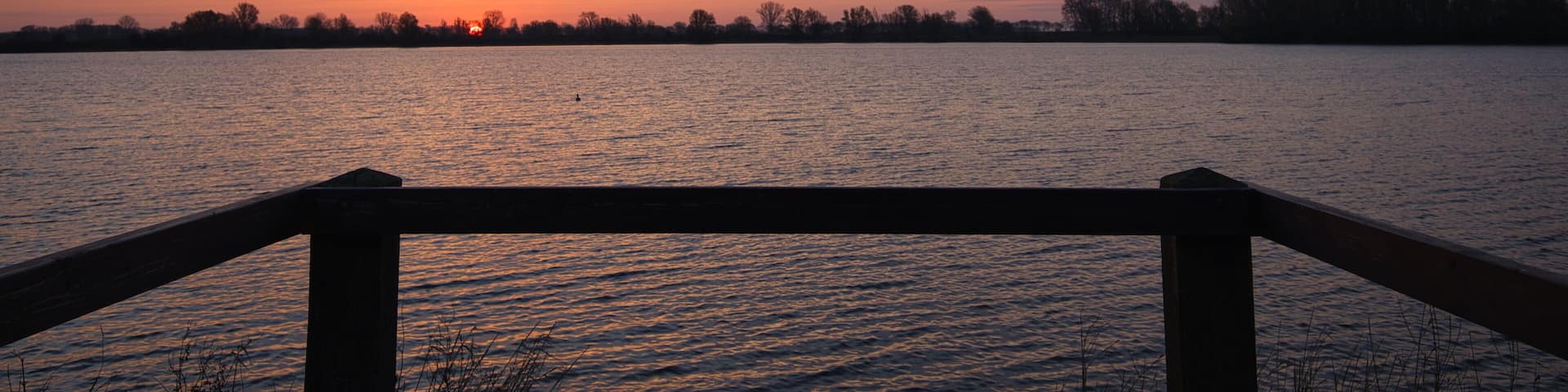 Horizontal view on a silhouette of a wooden railing with a beautiful sunrise, clouds and water at the background. Early morning in spring at “De Zandmeren” in Kerkdriel, Netherlands