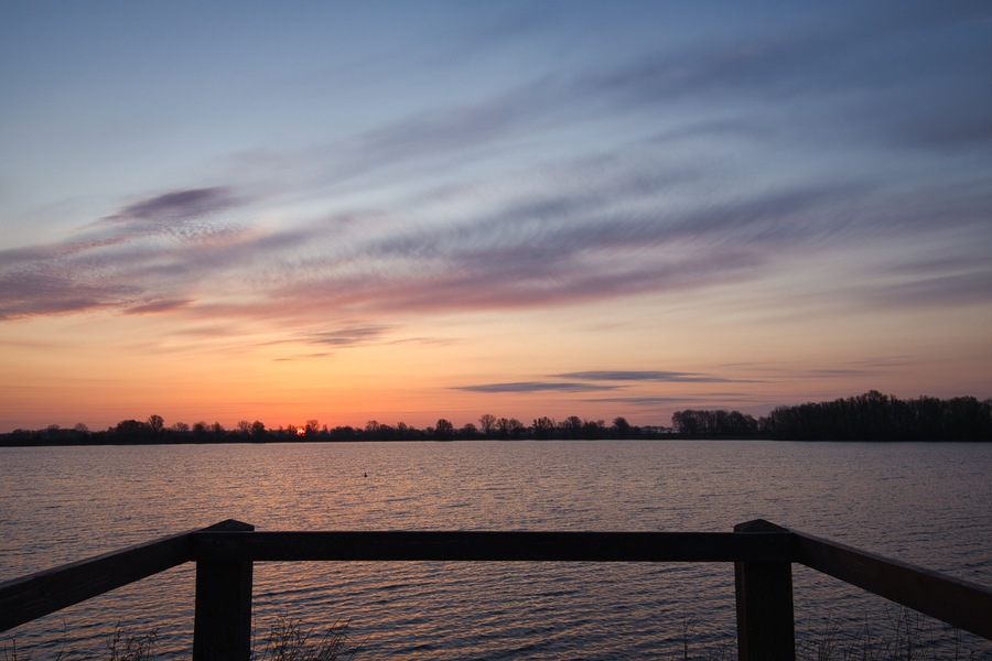 Horizontal view on a silhouette of a wooden railing with a beautiful sunrise, clouds and water at the background. Early morning in spring at “De Zandmeren” in Kerkdriel, Netherlands