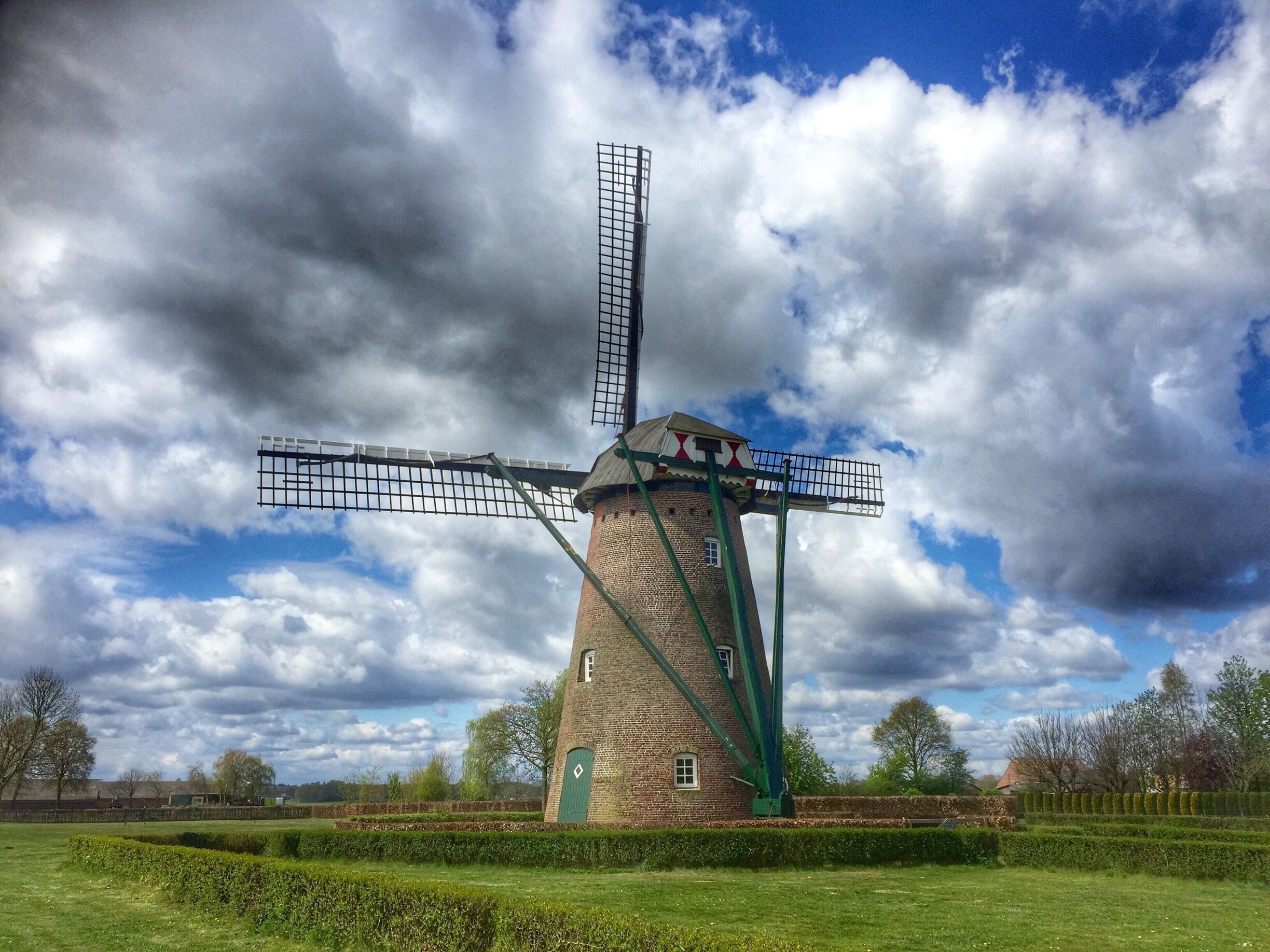 A traditional windmill with a large sail, surrounded by a well-maintained garden and trees under a partly cloudy sky.