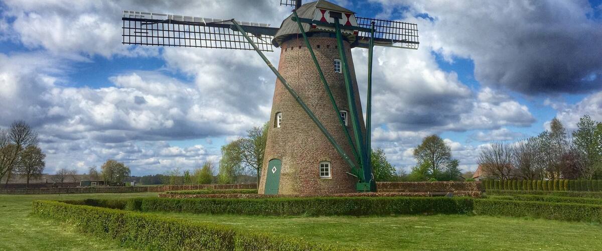 A traditional windmill with a large sail, surrounded by a well-maintained garden and trees under a partly cloudy sky.