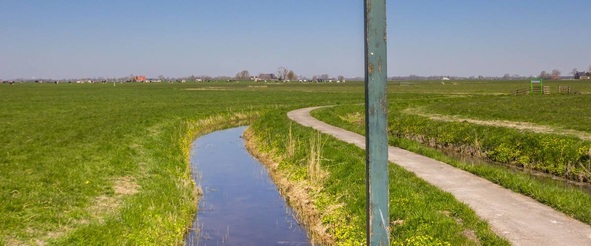 A rural landscape with a canal, green fields, and a signpost indicating directions to Den Ham and Fransum.