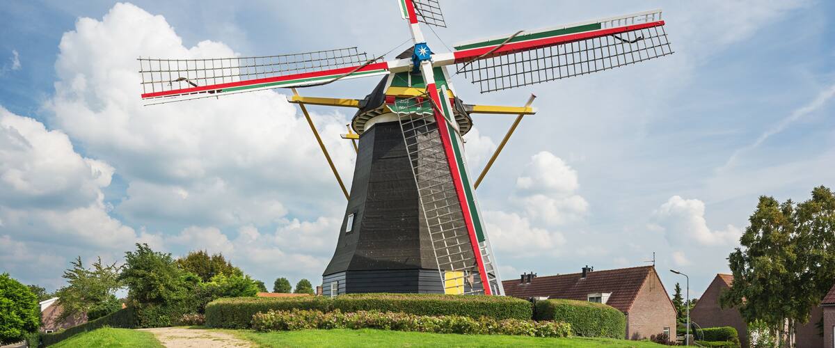 A traditional windmill with a black base, red and green sails, and a blue top, set against a partly cloudy sky.