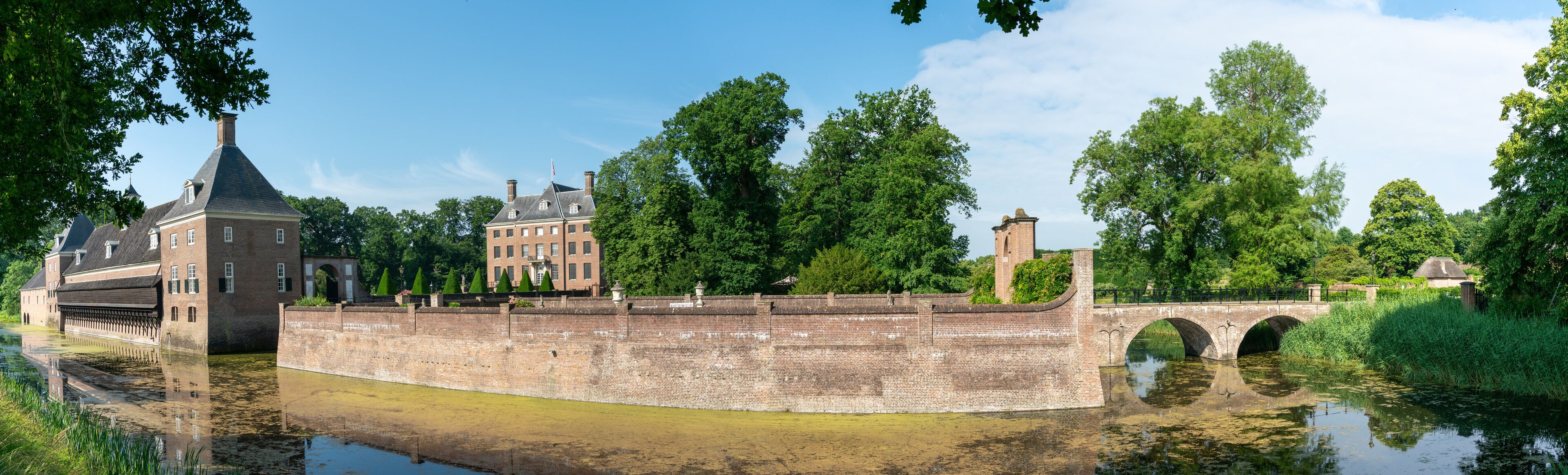 A historic brick bridge over a canal, surrounded by lush greenery and a prominent red brick building with a tower.