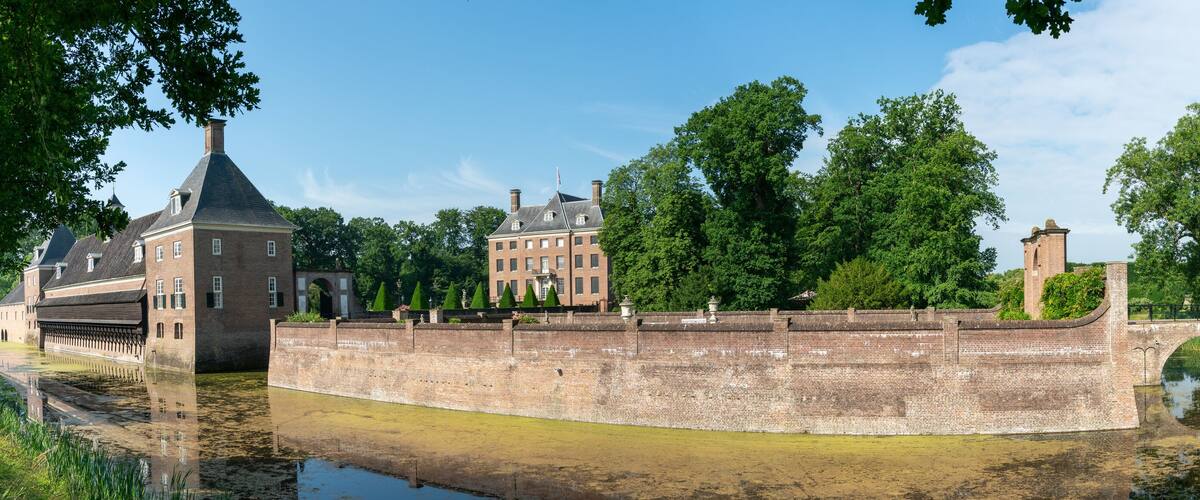 A historic brick bridge over a canal, surrounded by lush greenery and a prominent red brick building with a tower.