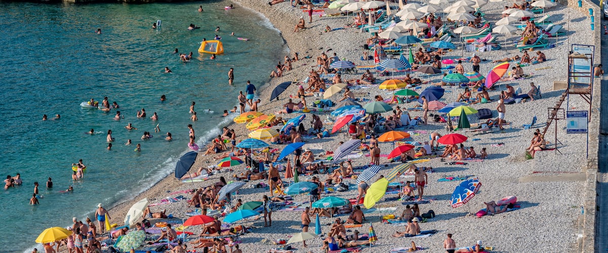 A crowded beach with people sunbathing and swimming, surrounded by colorful umbrellas.