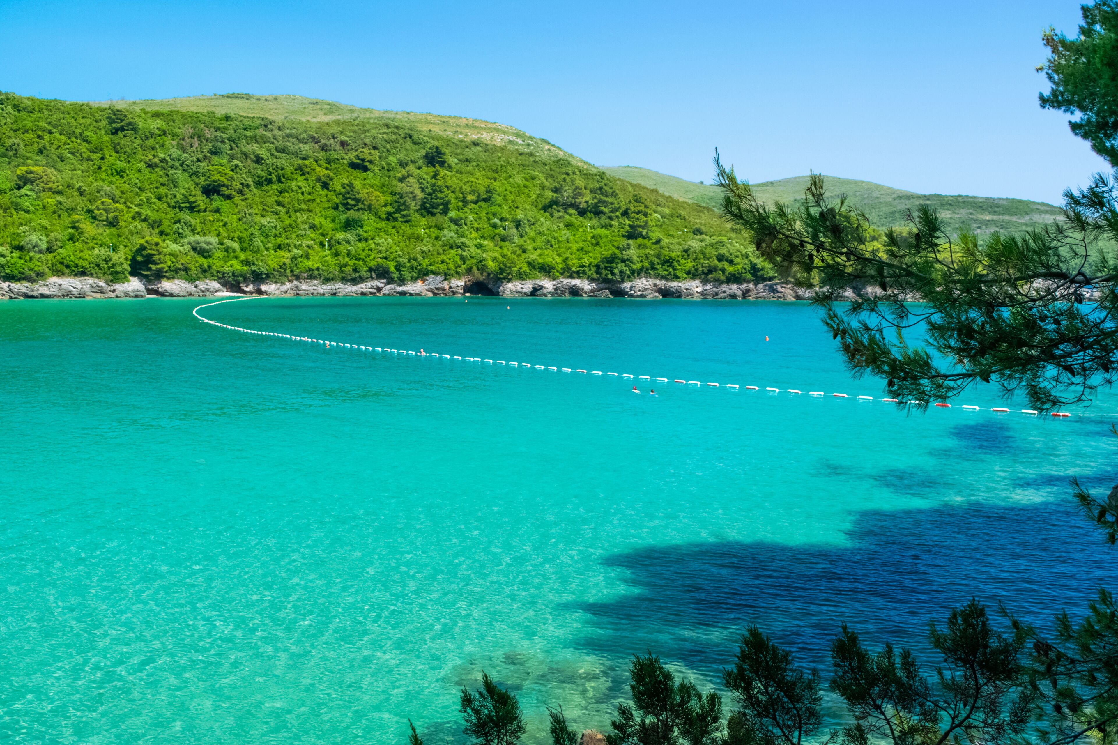 A turquoise sea with a white rope barrier, surrounded by green hills and pine trees.