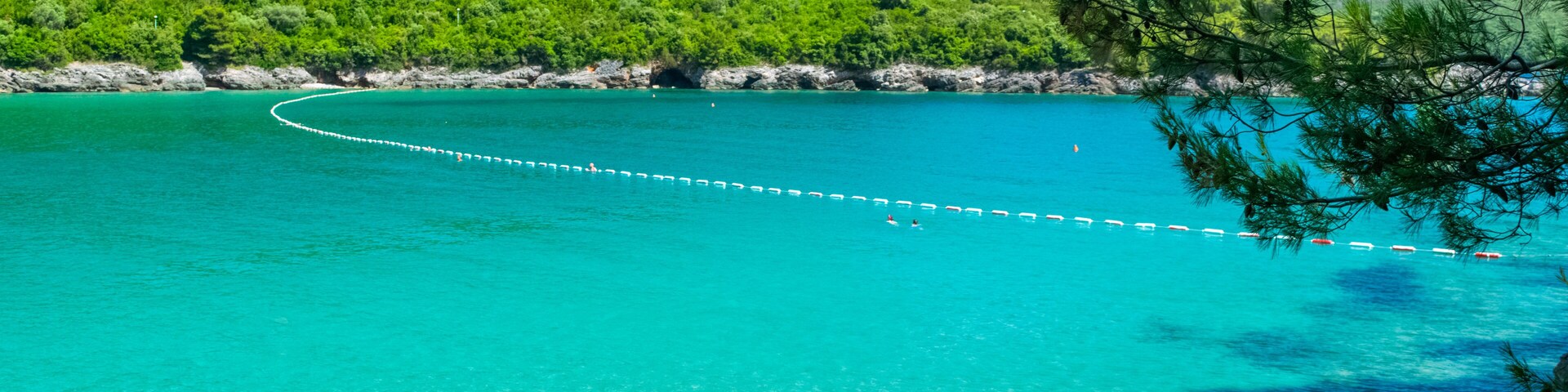 A turquoise sea with a white rope barrier, surrounded by green hills and pine trees.