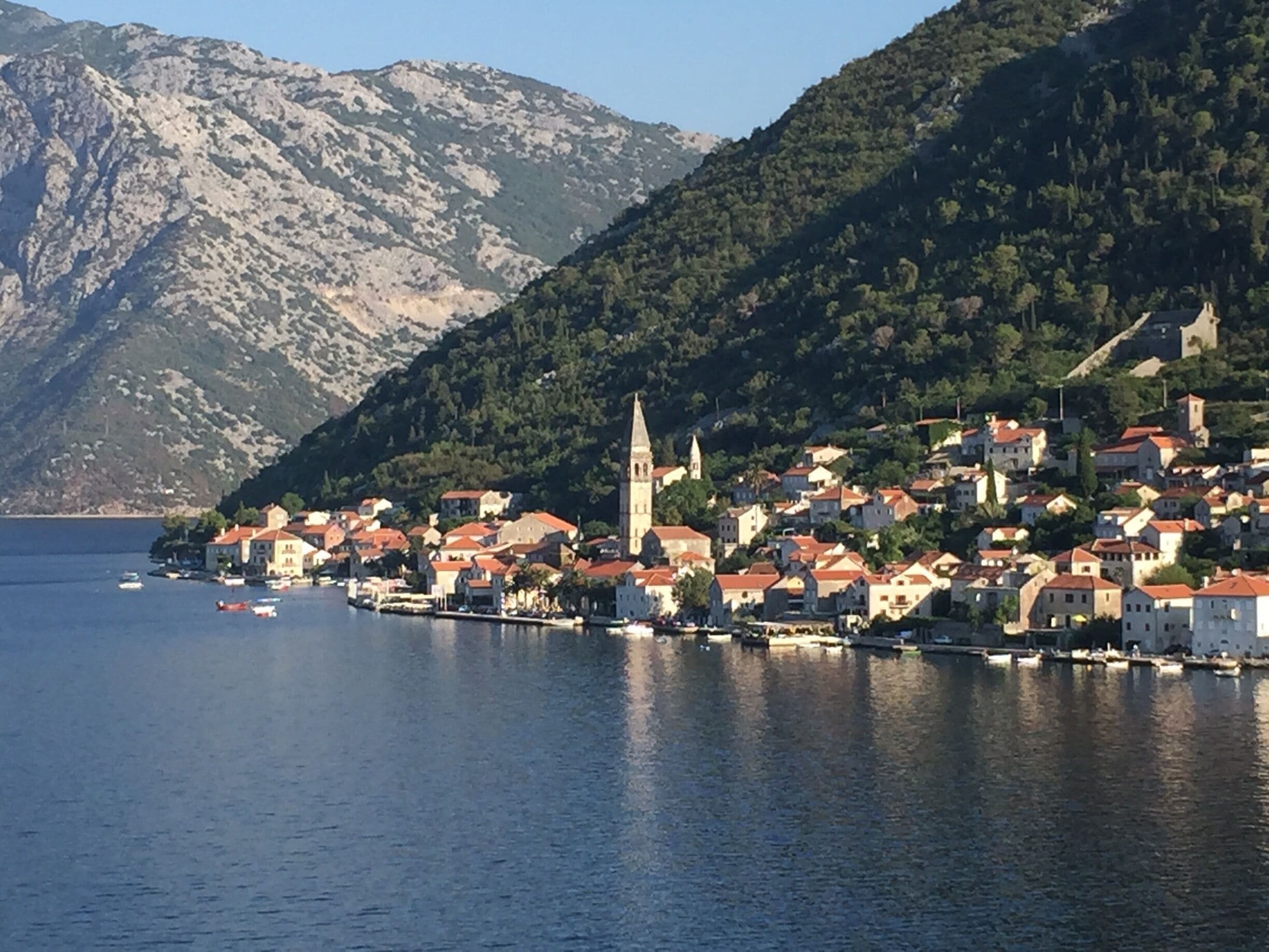 A coastal town with red-roofed buildings, a prominent church tower, and a mountainous backdrop.
