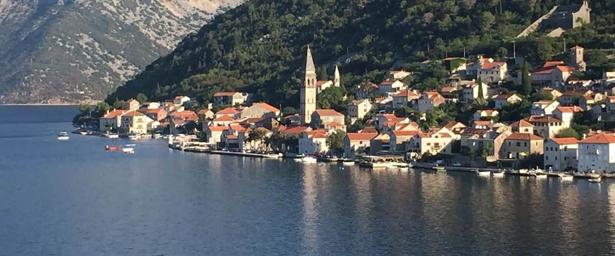 A coastal town with red-roofed buildings, a prominent church tower, and a mountainous backdrop.