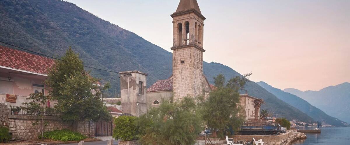 A stone pier with two boats docked, a church with a bell tower, and a mountainous backdrop.