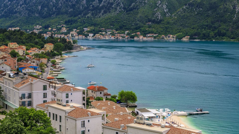 A coastal town with terracotta-roofed buildings, a marina, and a backdrop of green hills.