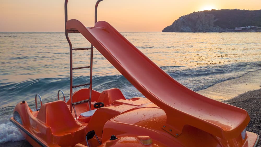 Slide on orange pedal (paddle) boat - pedalo parked on a sandy beach at sunset. Calm blue sea water and clear pink sky in the background. In Canj (Čanj) Montenegro.