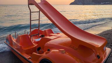 Slide on orange pedal (paddle) boat - pedalo parked on a sandy beach at sunset. Calm blue sea water and clear pink sky in the background. In Canj (Čanj) Montenegro.