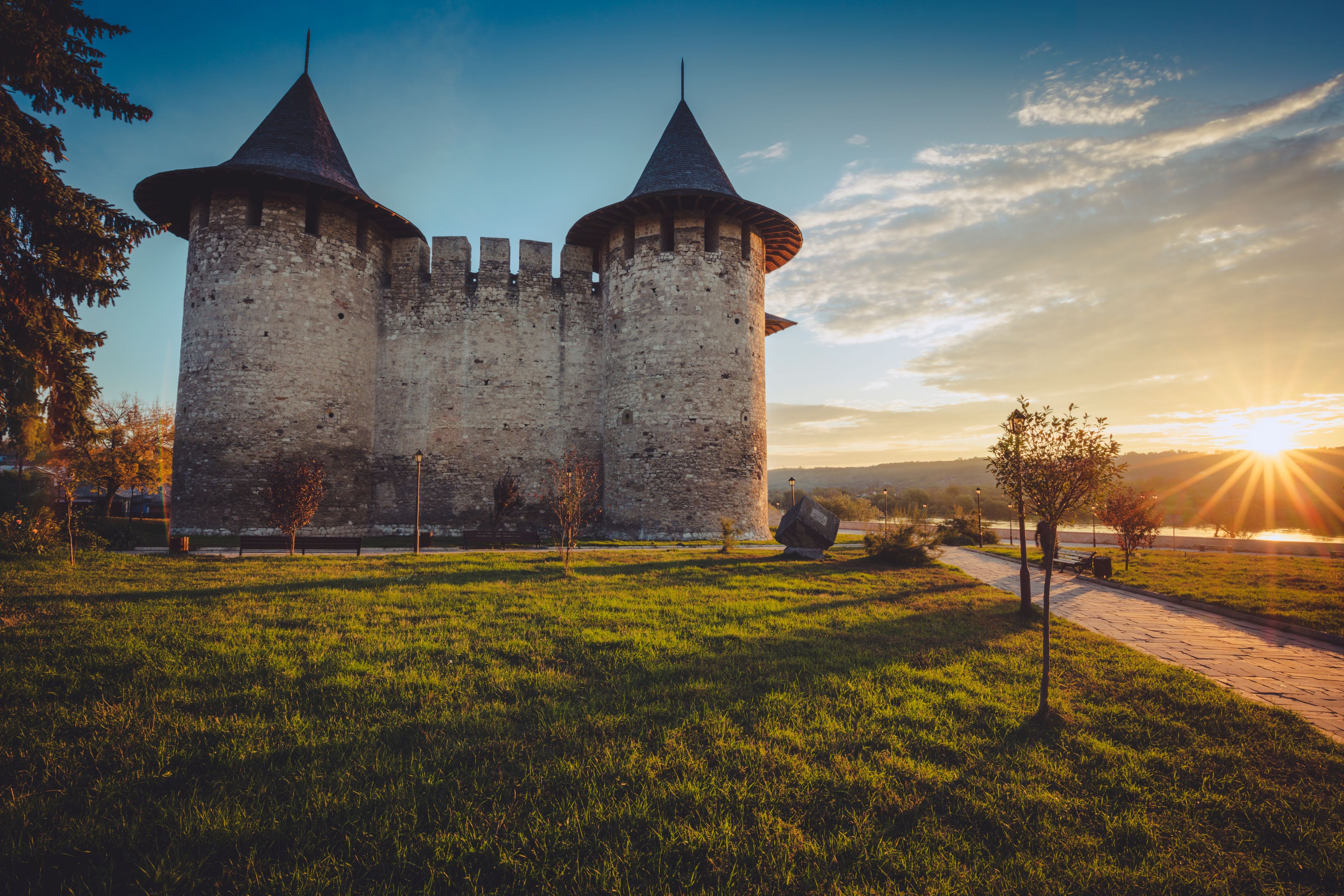 A medieval castle with two towers, surrounded by a grassy field and a pathway leading to a river.