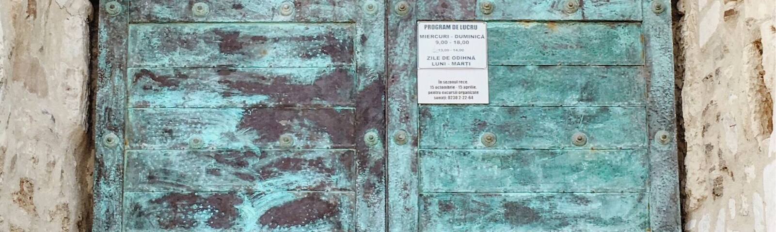 Mulher sentada em frente a uma porta grande e desgastada, de cor turquesa, com uma placa.