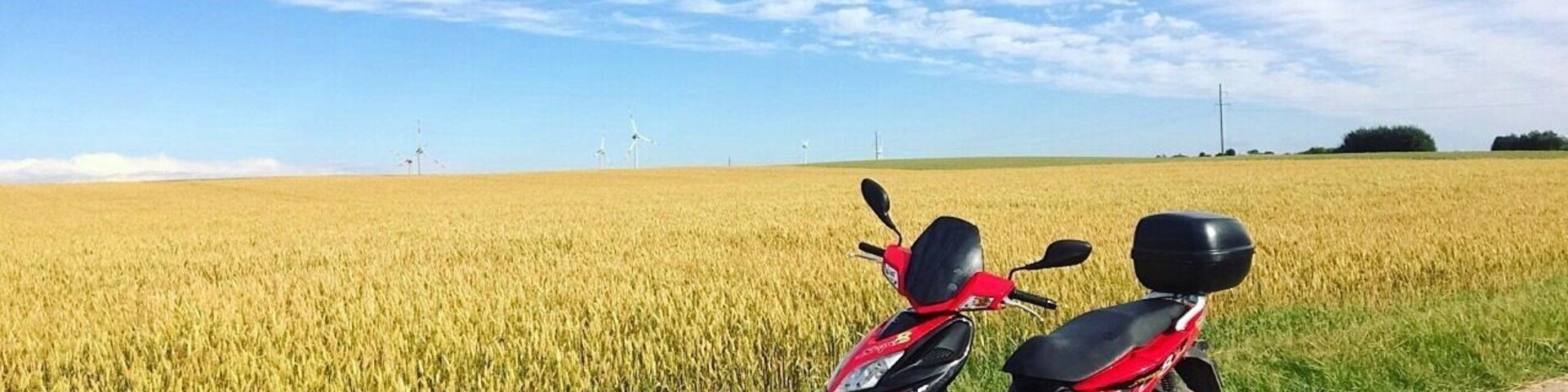 Duas motocicletas vermelhas estacionadas em uma trilha de terra ao lado de um campo dourado de trigo sob um céu parcialmente nublado.