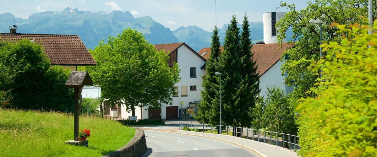 A curved road in a village with houses, trees, and mountains in the background.