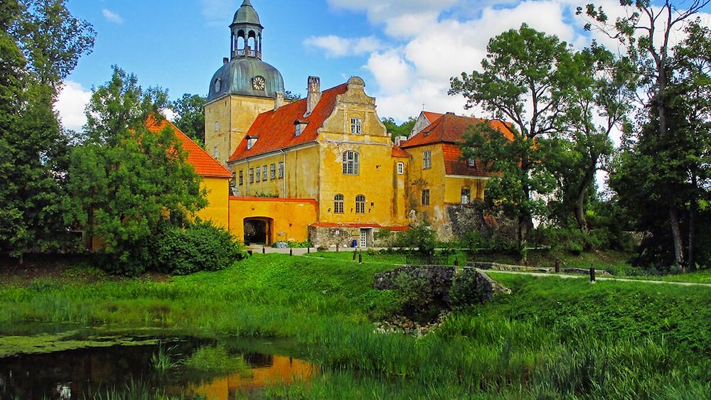 Un bâtiment jaune d’époque, doté d’un clocher, entouré d’une végétation luxuriante et bordé d’un étang.