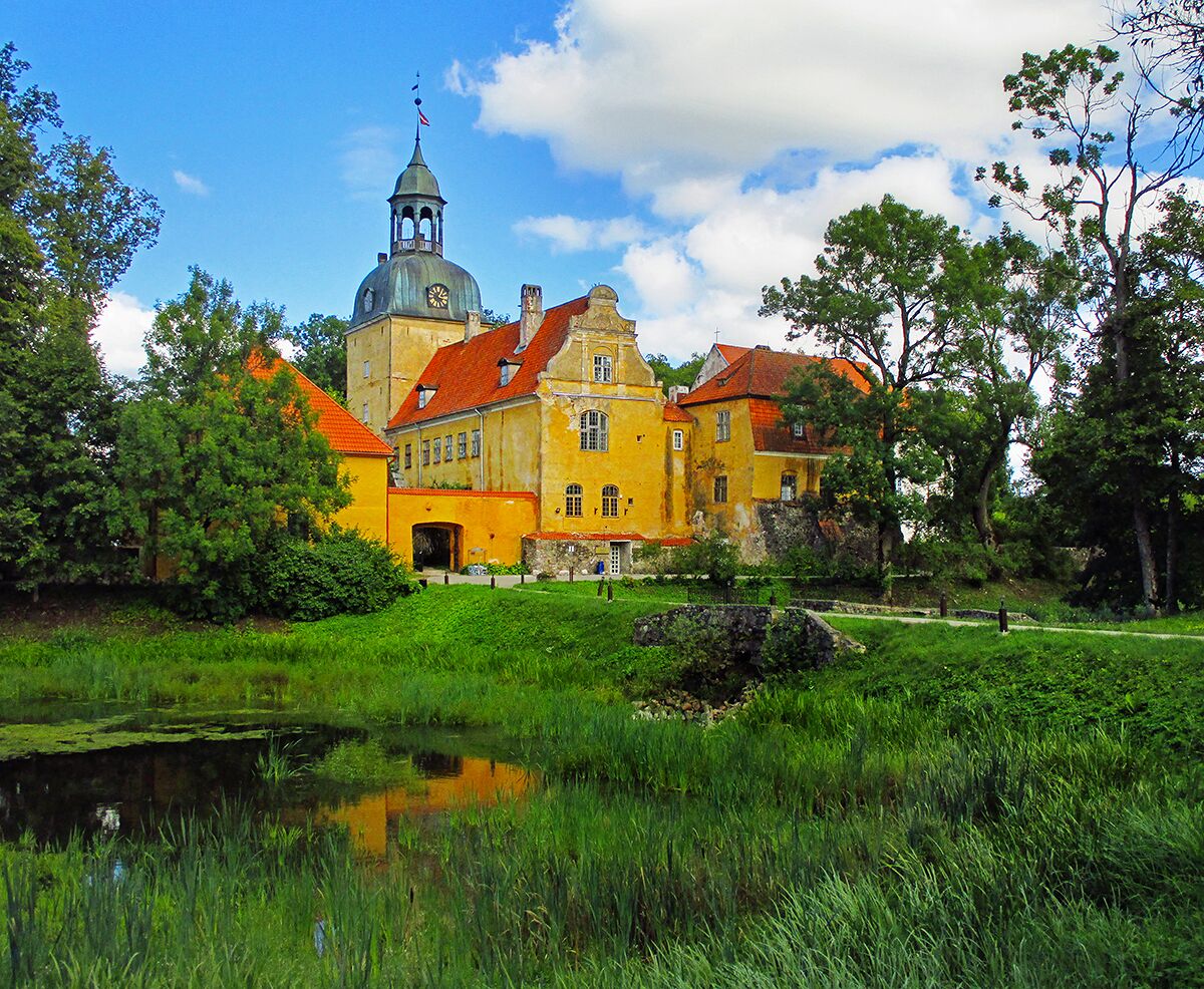 A historic yellow building with a clock tower, surrounded by lush greenery and a pond.