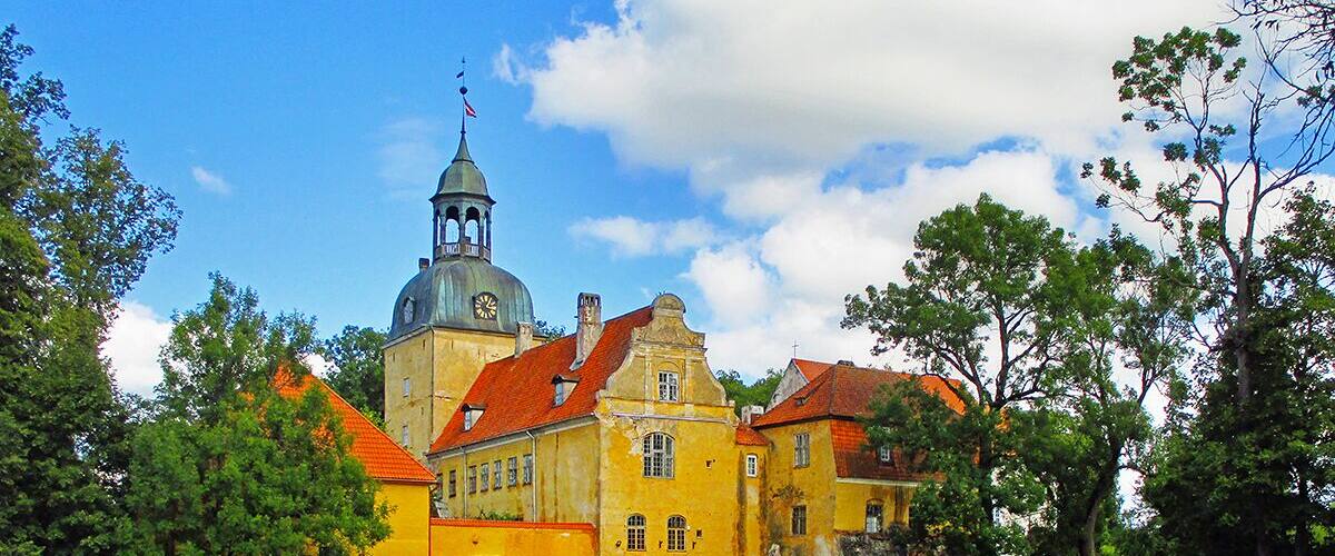 A historic yellow building with a clock tower, surrounded by lush greenery and a pond.