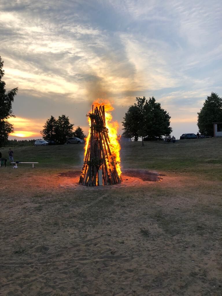 A bonfire in a grassy field with trees and a sunset in the background.