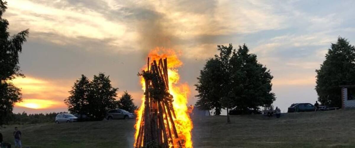 A bonfire in a grassy field with trees and a sunset in the background.