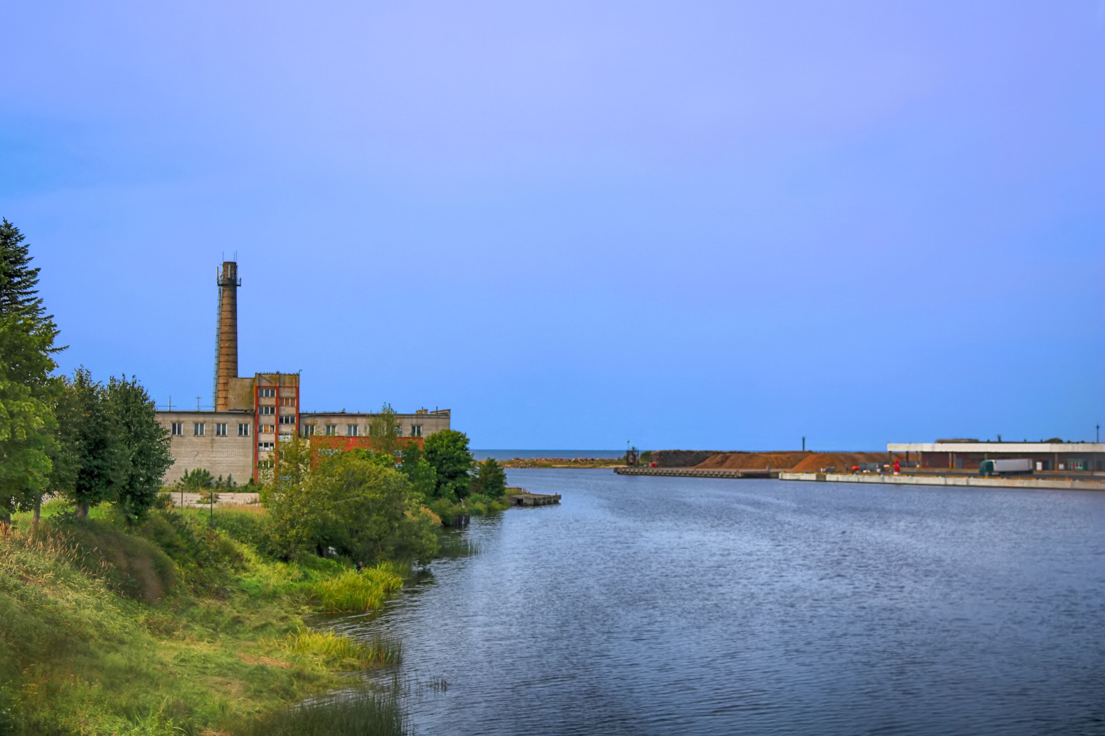 A river with a factory and a tall chimney on the left, surrounded by greenery and a clear sky.