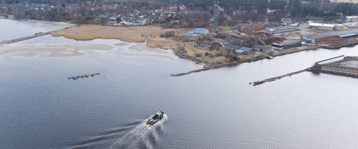 A boat moving across a calm body of water with a shoreline featuring industrial buildings and a bridge.