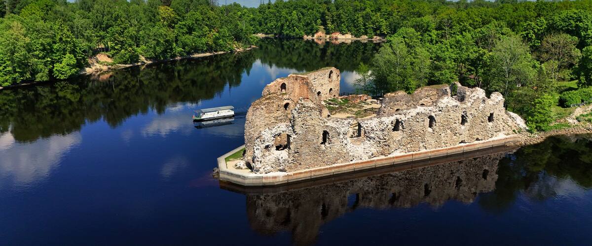 Eine historische Steinburg, umgeben von einem stillen See, mit dichtem, grünen Wald im Hintergrund.