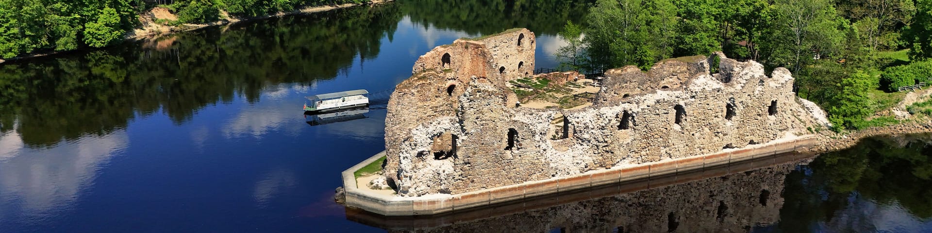 A historic stone fortress surrounded by a calm lake, with dense green forest in the background.