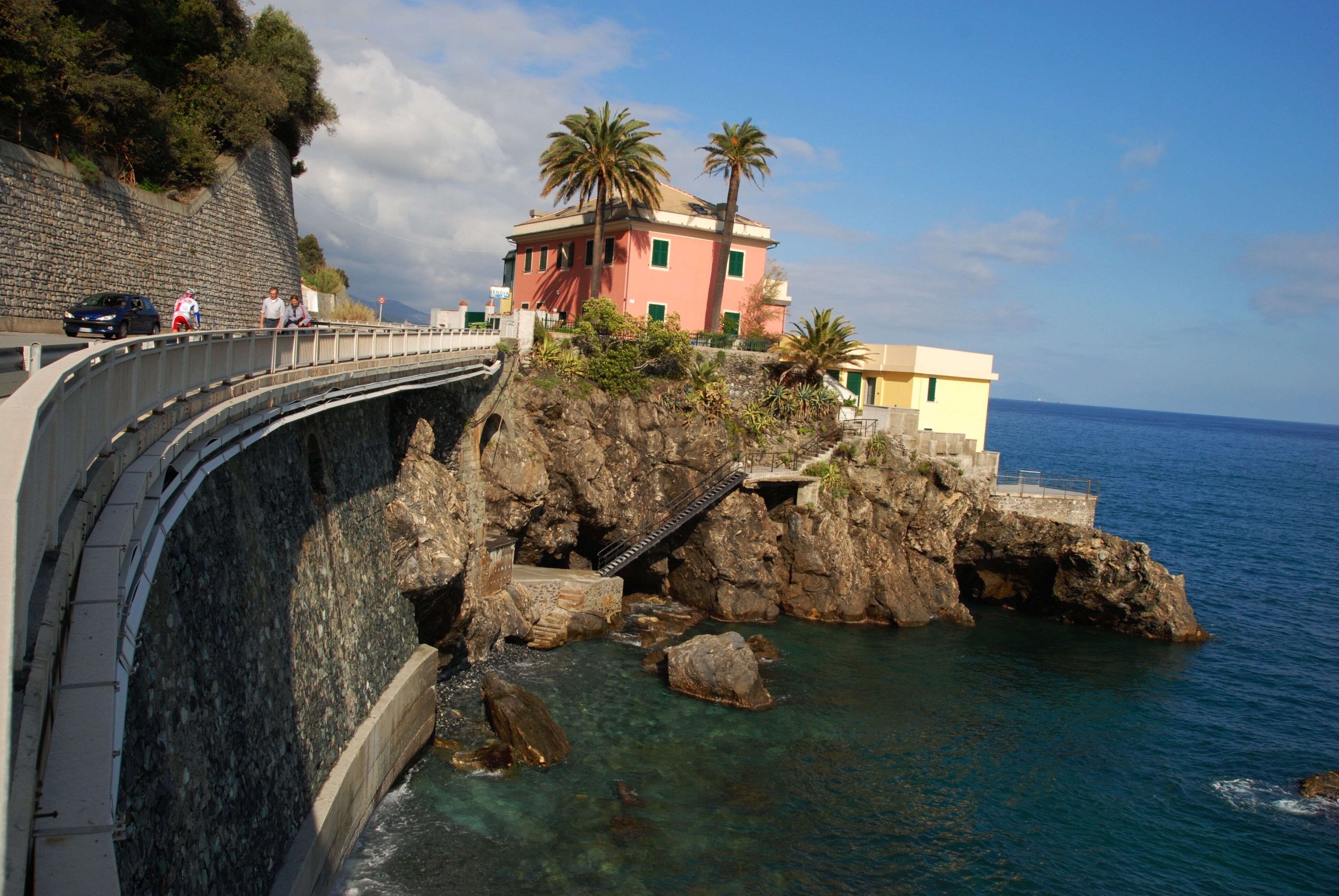 A coastal road with a bridge, rocky cliffs, and a building with palm trees.