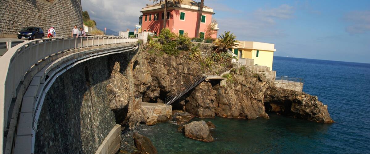 A coastal road with a bridge, rocky cliffs, and a building with palm trees.