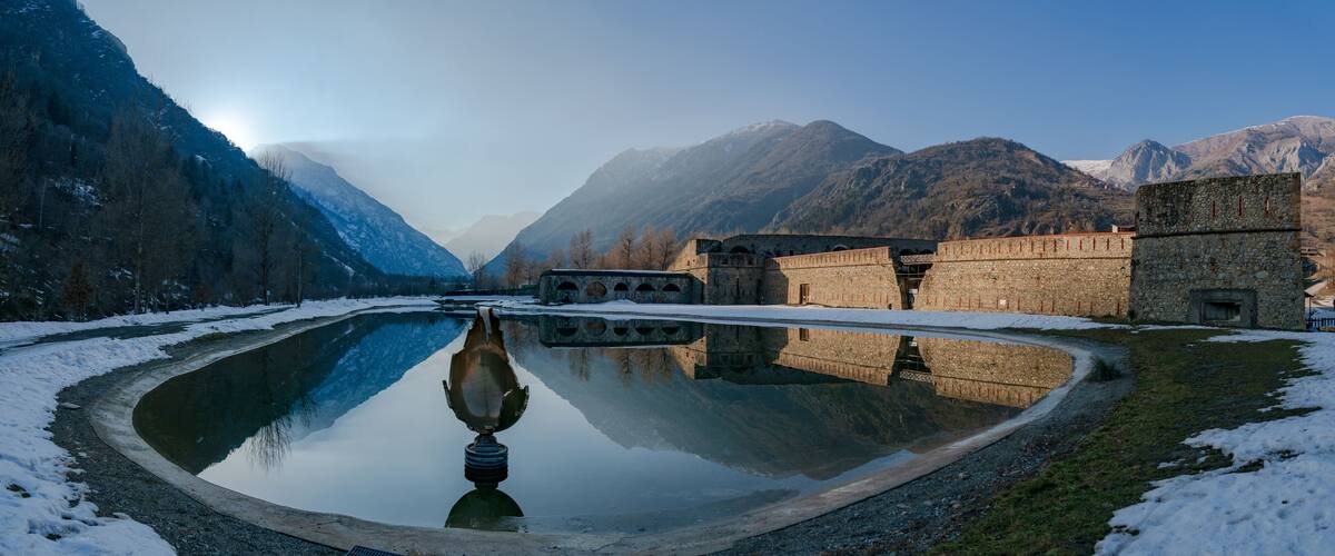 A snow-covered landscape with a reflective pond, stone buildings, and mountains in the background.