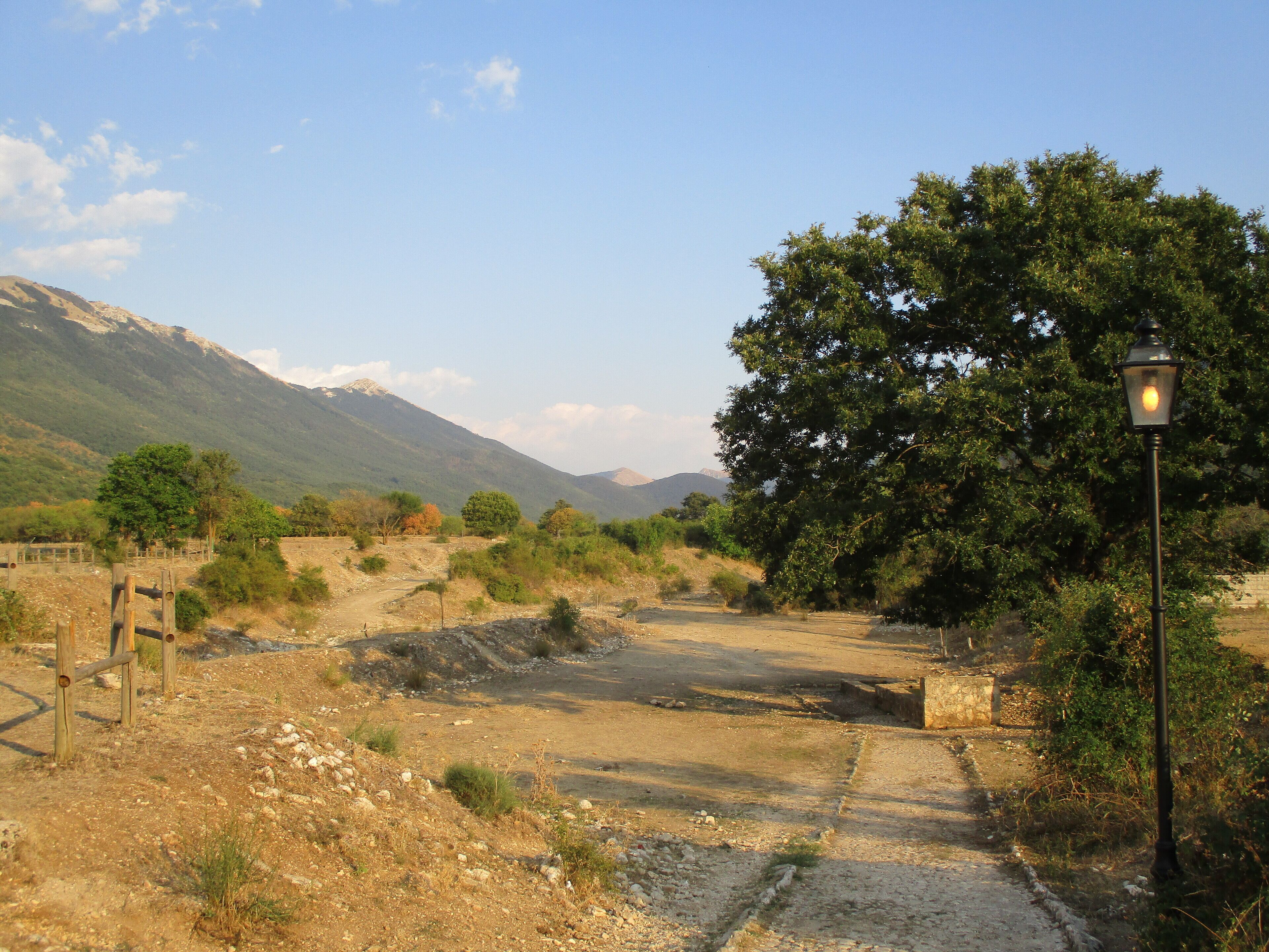 A dirt path leading through a rural landscape with a large tree on the right and mountains in the background.