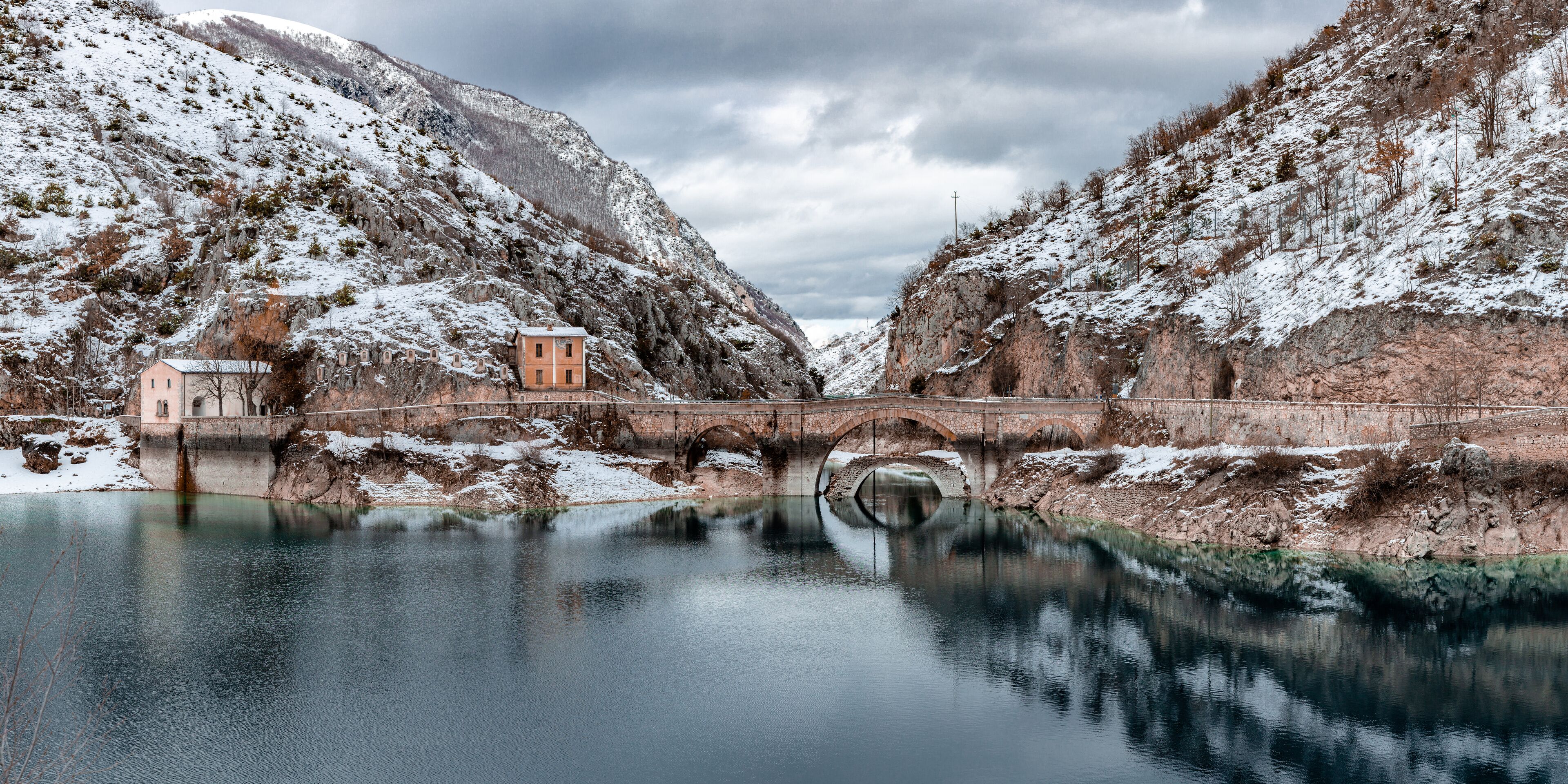 A snow-covered landscape with a bridge over a calm river, reflecting the surrounding scenery.