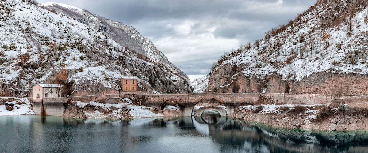 A snow-covered landscape with a bridge over a calm river, reflecting the surrounding scenery.
